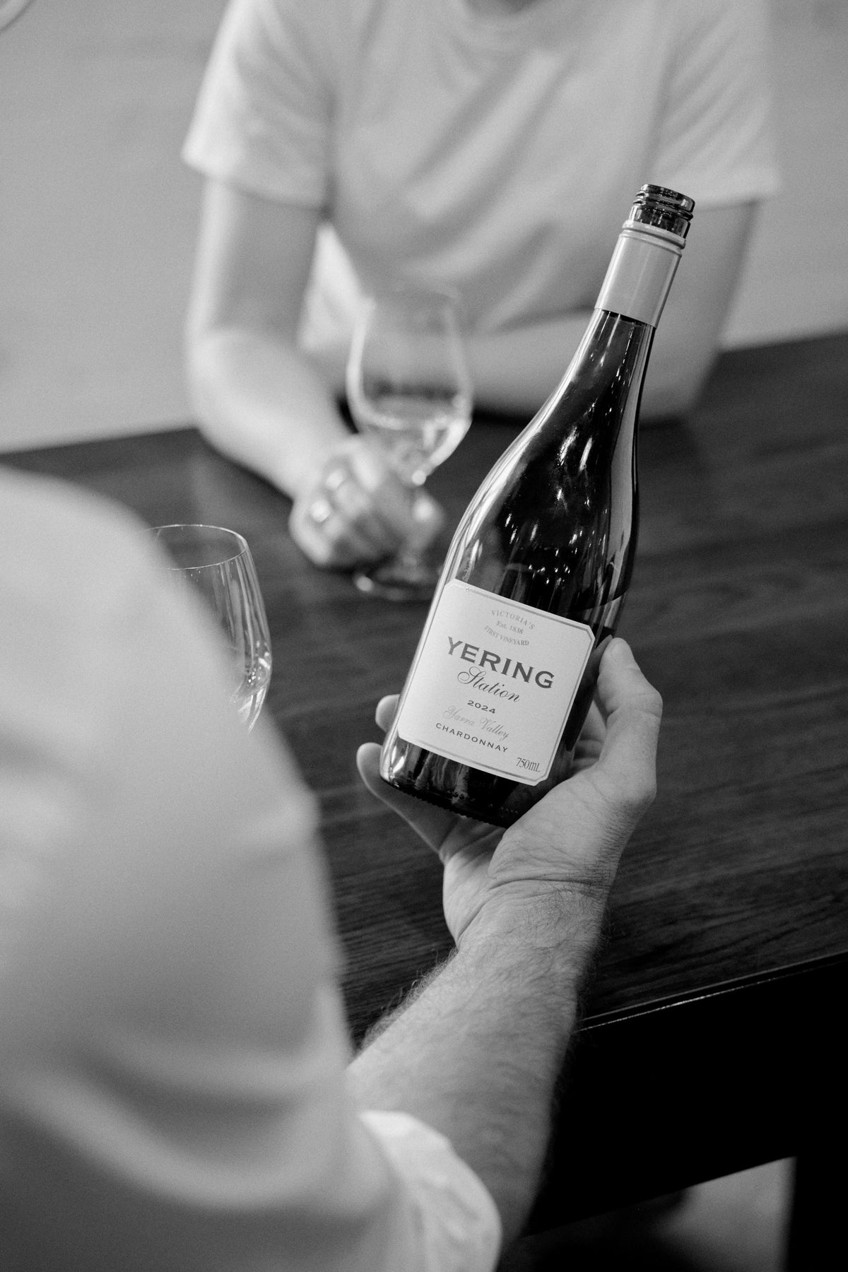 Black and white photo of a host presenting a bottle of Yering Station wine to a guest at a tasting table, with wine glasses in the foreground