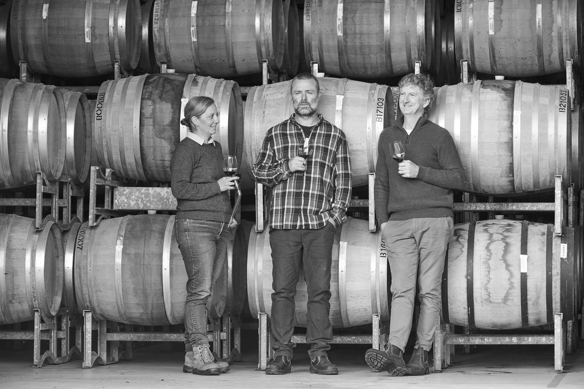 Black and white photo of three winemakers standing among wine barrels, each holding a glass of red wine