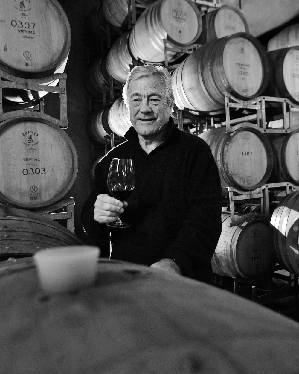 Black and white portrait of a man holding a glass of red wine among wine barrels in the cellar