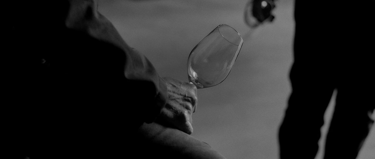Black and white close-up of a hand swirling a wine glass with a bottle in the background