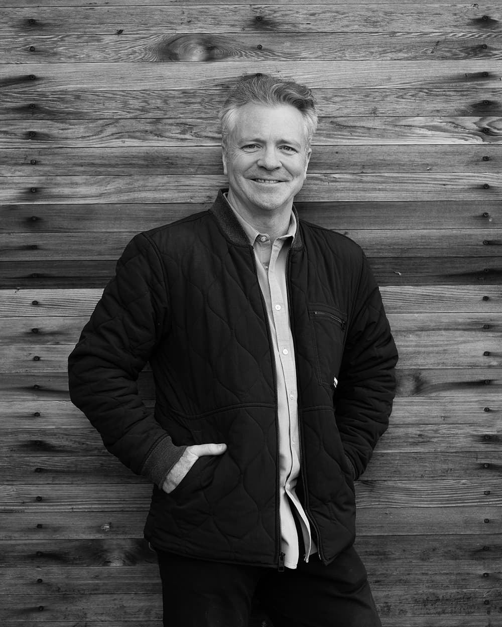 Black and white portrait of a smiling man in a quilted jacket standing against a timber wall