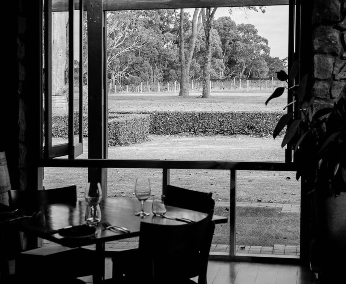 Black and white view through a restaurant window overlooking a lawn and vineyard, set dining table in foreground