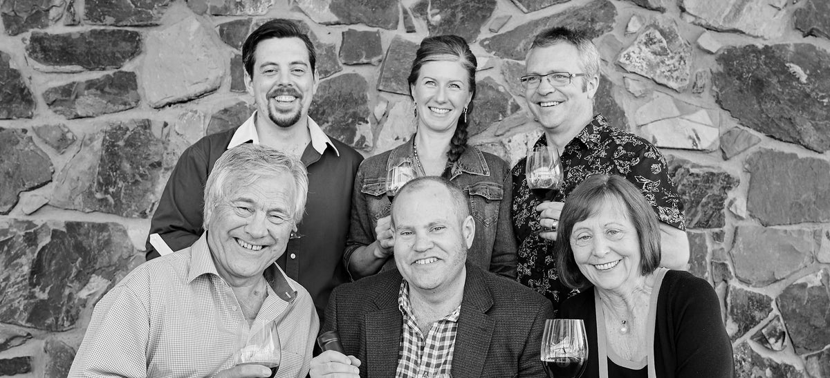Black and white group photo of the Rathbone family and winemaking team smiling in front of a stone wall, holding wine glasses