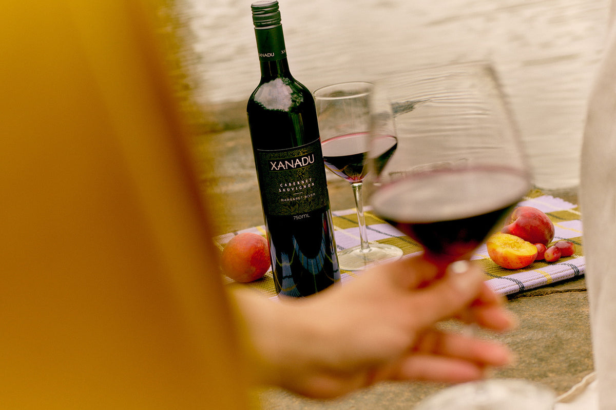 Person holding a glass of red wine beside a bottle of Xanadu Cabernet Sauvignon, with peaches and grapes on a picnic mat
