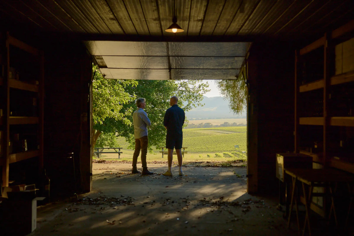 Two people standing in a winery shed doorway looking out over the vineyard at sunrise