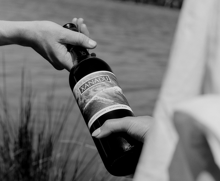 Black and white close-up of hands holding a bottle of Xanadu wine outdoors by the water