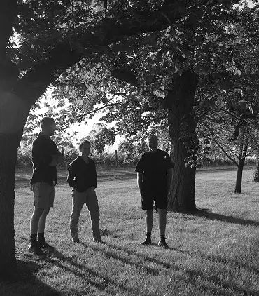 Black and white photo of three men standing together under a large tree in a vineyard