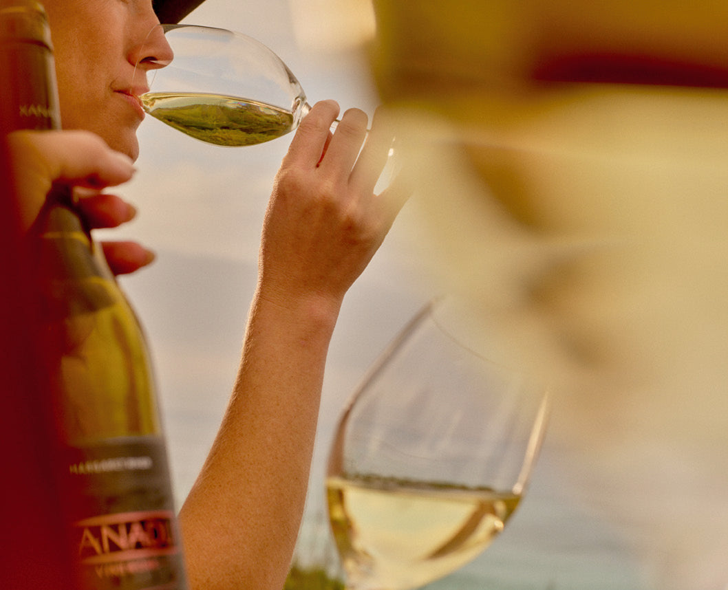 Close-up of a person tasting white wine with a bottle and glass by the water, Xanadu wine club banner
